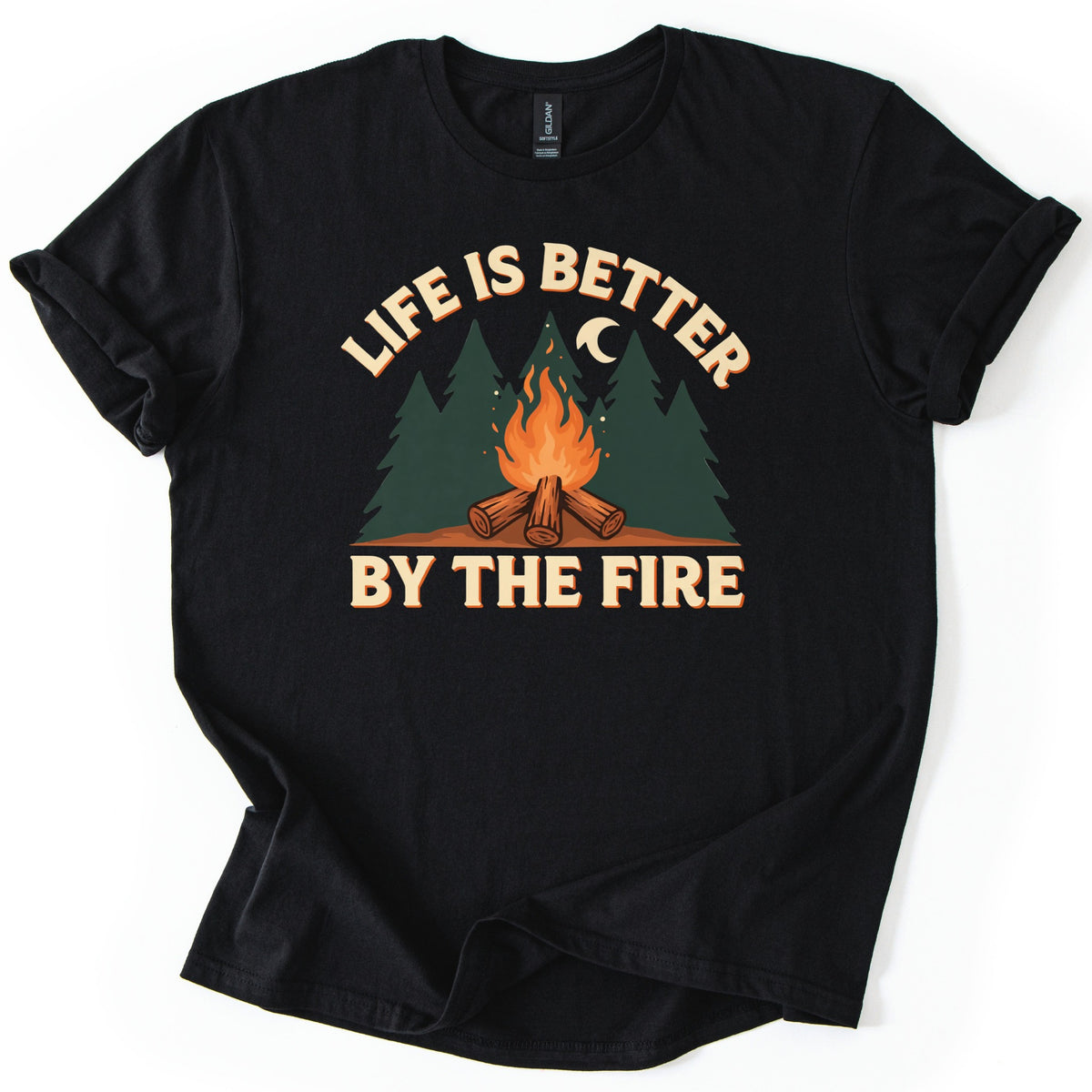 Life Is Better By The Fire Comfort Colors T-shirt in Black, laid flat. A campfire of crossed logs burns in front of dark green pine trees under a crescent moon, with LIFE IS BETTER arched above and BY THE FIRE below in cream letters. Cuffed sleeves, white background.