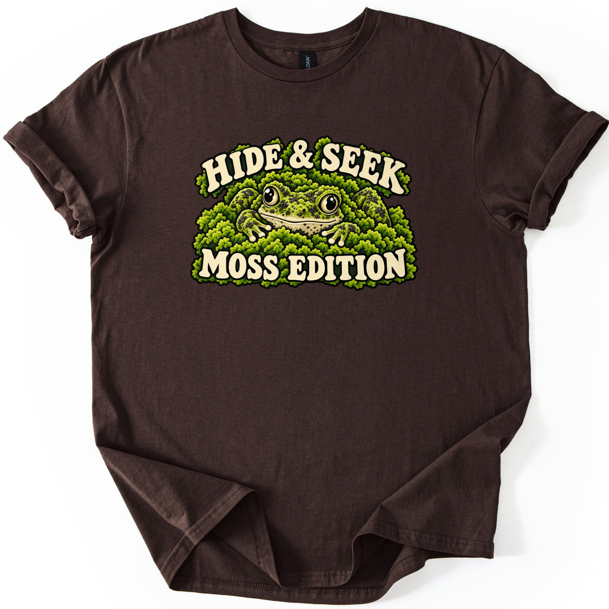 Hide & Seek Moss Edition Comfort Colors T-shirt in Dark Chocolate, laid flat. A green speckled frog with large eyes nestles in bright green moss, with Hide & Seek arched above and Moss Edition below in cream letters. The shirt lies on white with rolled sleeves.