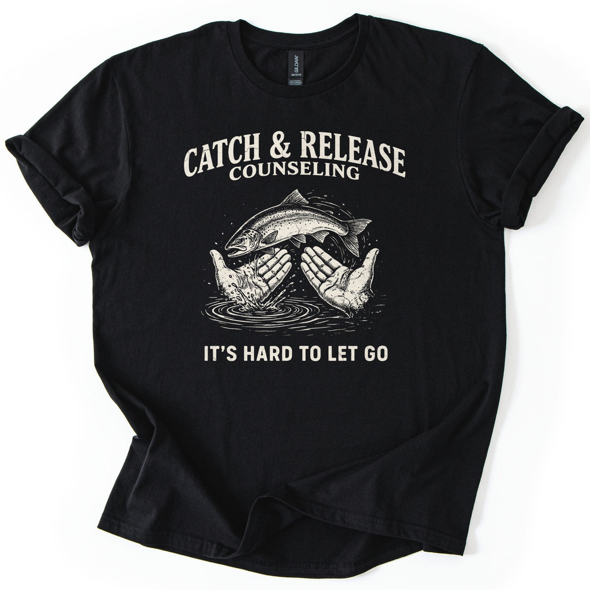 Catch & Release Counseling Comfort Colors T-shirt in Black, laid flat. Cream print reads CATCH & RELEASE COUNSELING above an etched trout leaping from two open hands over rippling water, with IT'S HARD TO LET GO below. Sleeves are cuffed on a white background.
