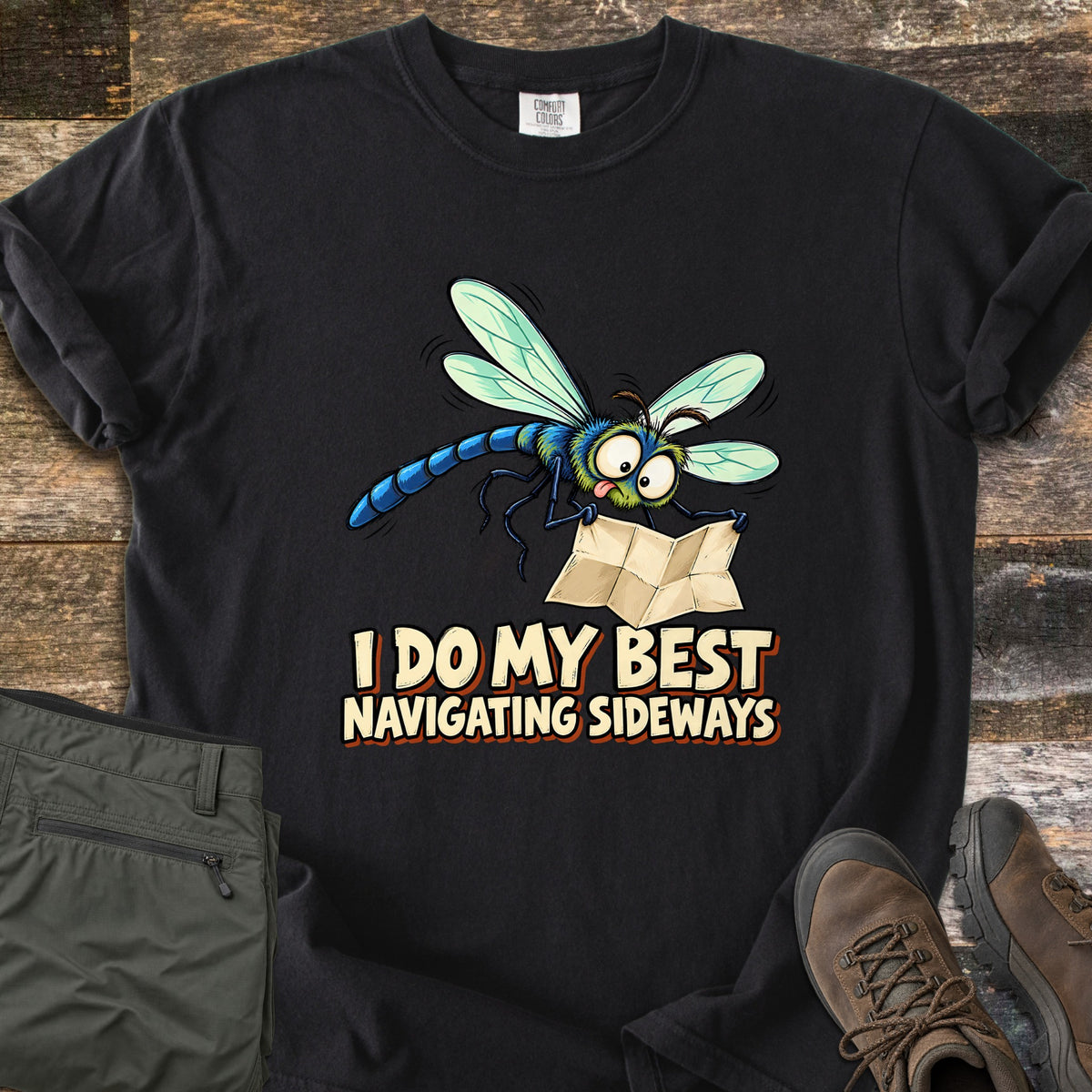 I Do My Best Navigating Sideways Comfort Colors T-shirt in Black, laid flat. A wide-eyed cartoon dragonfly with its tongue out holds a folded paper map above the words I Do My Best Navigating Sideways. Olive pants and brown hiking boots lie beside it on weathered wood planks.