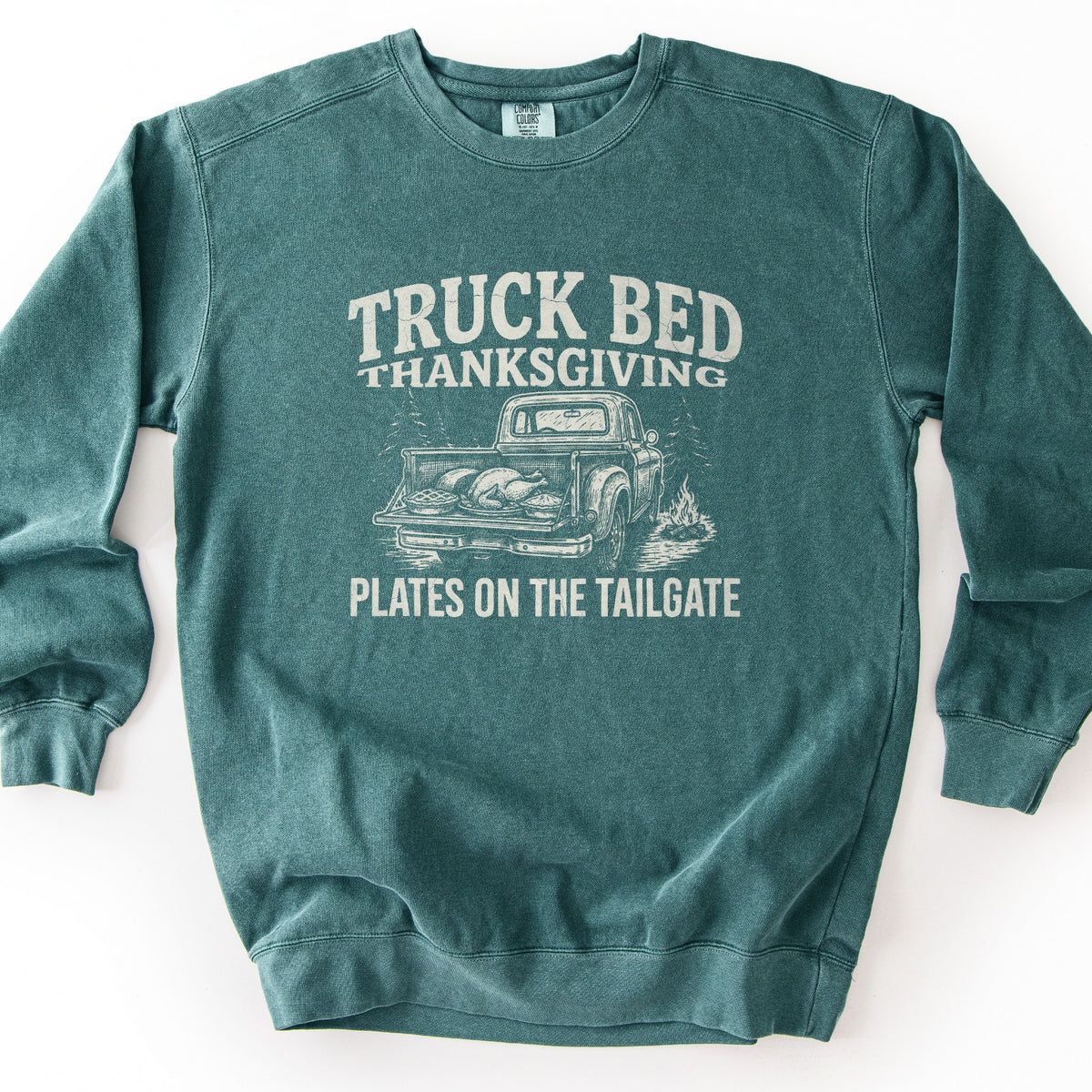 Truck Bed Thanksgiving Comfort Colors crewneck sweatshirt in Blue Spruce, laid flat. Distressed off-white print reads Truck Bed Thanksgiving above a sketched vintage pickup with a roast turkey and pies in its open bed, pine trees and a campfire, and the words Plates on the Tailgate. Shown on a white background.