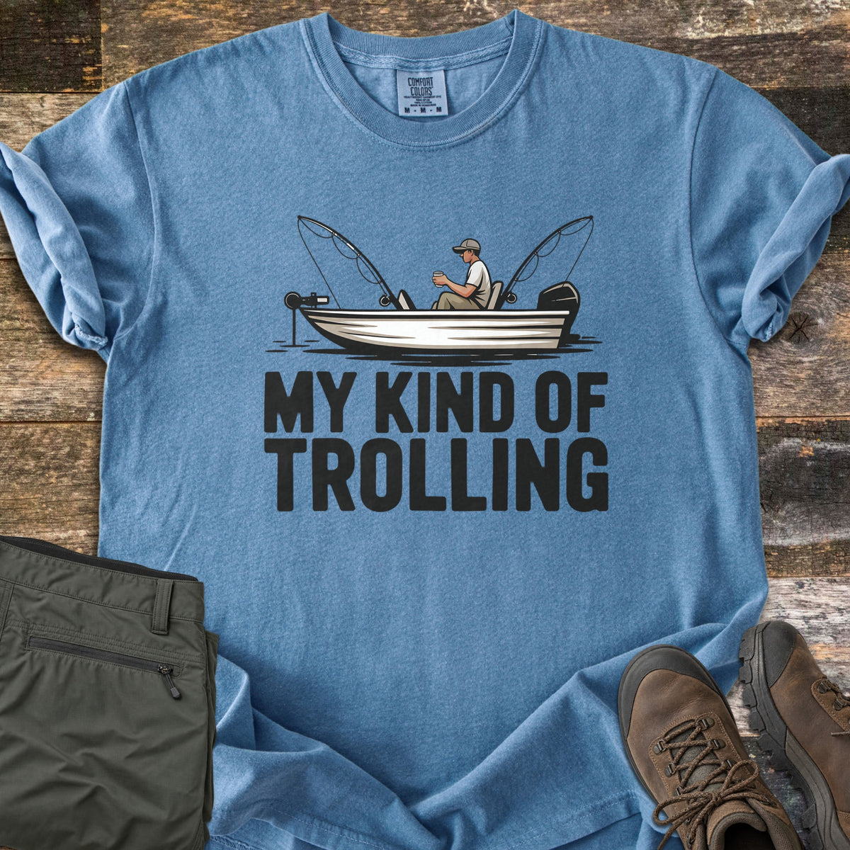 My Kind Of Trolling Comfort Colors T-shirt in Blue Jean, laid flat. A man in a cap holds a cup in a small fishing boat with two bent rods and motors, above the words My Kind Of Trolling in black. Olive cargo pants and brown hiking boots sit beside it on weathered wood.