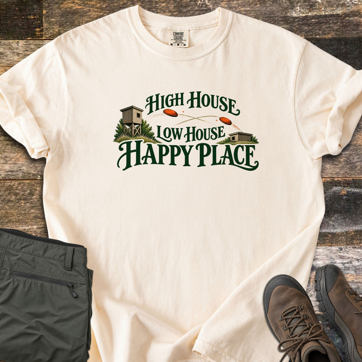High House Low House Happy Place Comfort Colors T-shirt in Ivory, laid flat. Green lettering reads High House Low House Happy Place around two skeet houses with orange clay targets flying between them. Olive cargo pants and brown hiking boots lie beside it on weathered wood planks.