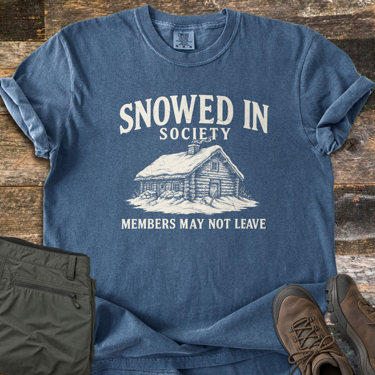 Snowed In Society Comfort Colors T-shirt in Denim, laid flat. A snow-covered log cabin with a smoking chimney sits between the words Snowed In Society and Members May Not Leave. It lies on weathered wood planks beside folded olive pants and brown hiking boots.