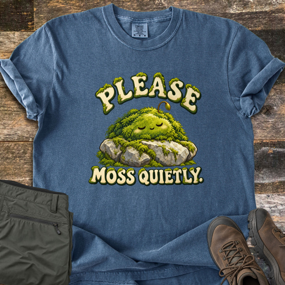 Please Moss Quietly Comfort Colors T-shirt in Denim, laid flat. A sleepy moss creature with closed eyes and a small sprout rests on gray rocks between mossy cream letters reading Please Moss Quietly. Olive hiking pants and brown hiking boots sit beside it on weathered wood.