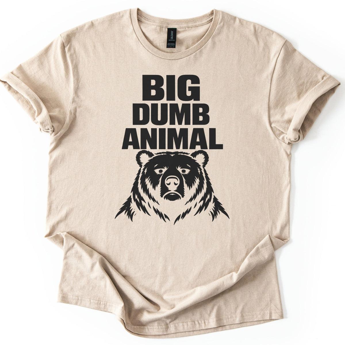 Big Dumb Animal Comfort Colors T-shirt in Sand, laid flat. Bold black letters read BIG DUMB ANIMAL above a black illustration of a stern, front-facing bear head with shaggy fur. The sleeves are rolled and the shirt lies on a white background.