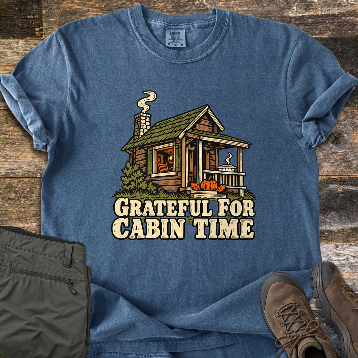 Grateful For Cabin Time Comfort Colors T-shirt in Denim, laid flat. A log cabin with a green roof, smoking stone chimney, glowing window, pie on the porch rail and a pumpkin on the steps, above the words Grateful For Cabin Time. Olive pants and brown hiking boots sit on weathered wood.