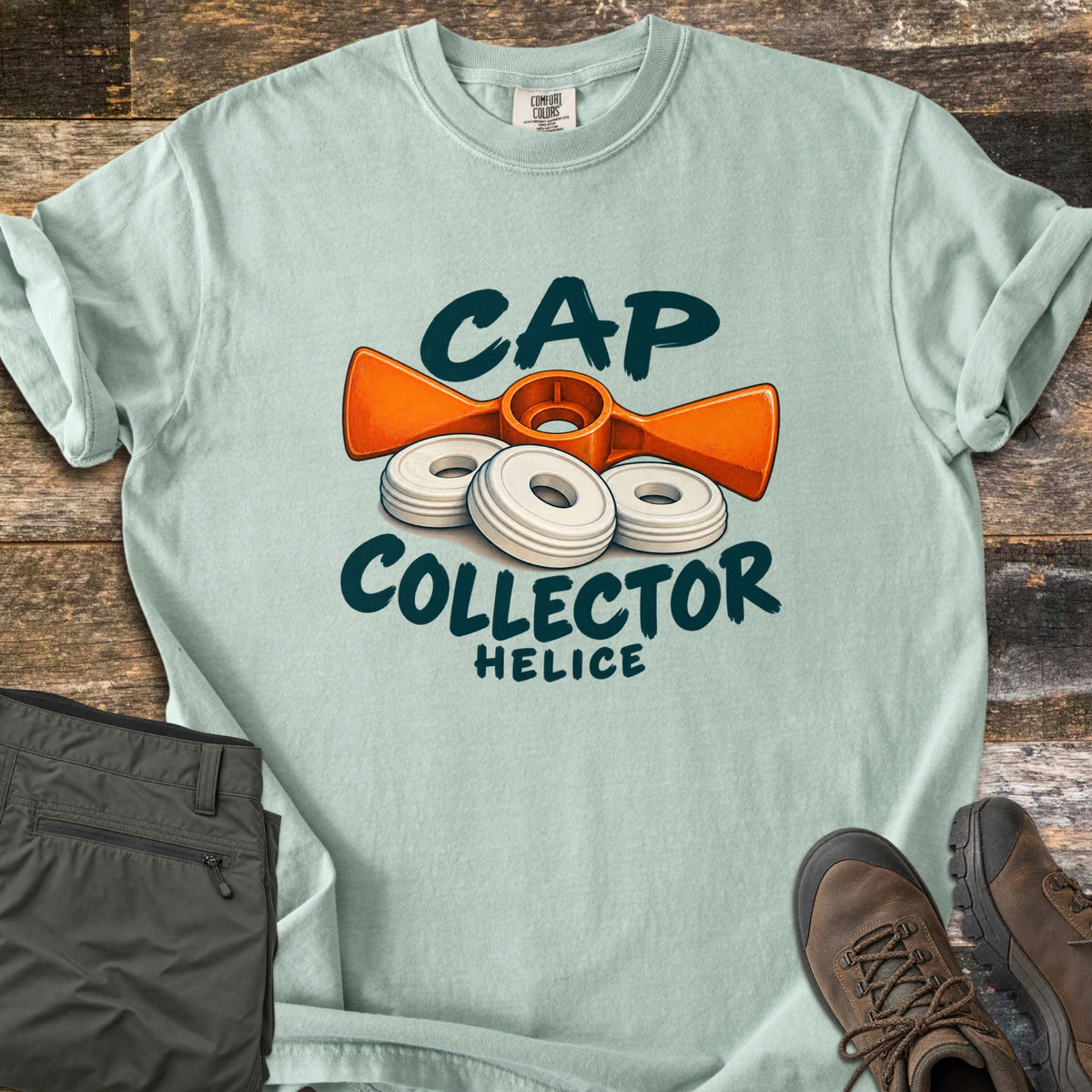 Cap Collector Comfort Colors T-shirt in Bay, laid flat. An orange helice key and three white caps sit between the words Cap Collector, with Helice below. Sleeves are rolled; olive pants and brown hiking boots lie beside it on dark wood planks.