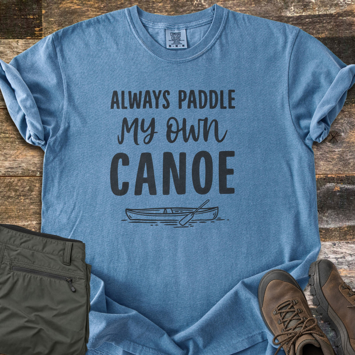 Paddle My Own Canoe Comfort Colors T-shirt in Blue Jean, laid flat. Charcoal text reads Always Paddle My Own Canoe above a line drawing of a canoe with a paddle on the water. It lies on weathered wood with rolled sleeves, folded olive hiking pants, and brown hiking boots.