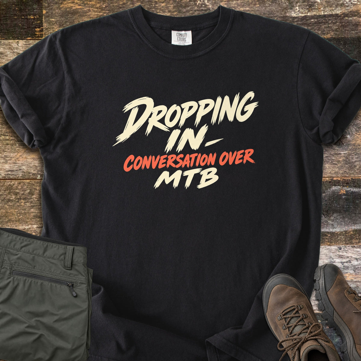 Dropping In Conversation Over Comfort Colors T-shirt in Black, laid flat. Cream brush-stroke letters read Dropping In- and MTB, with Conversation Over in orange-red between them. The shirt lies on dark weathered wood beside folded olive pants and brown leather hiking boots.