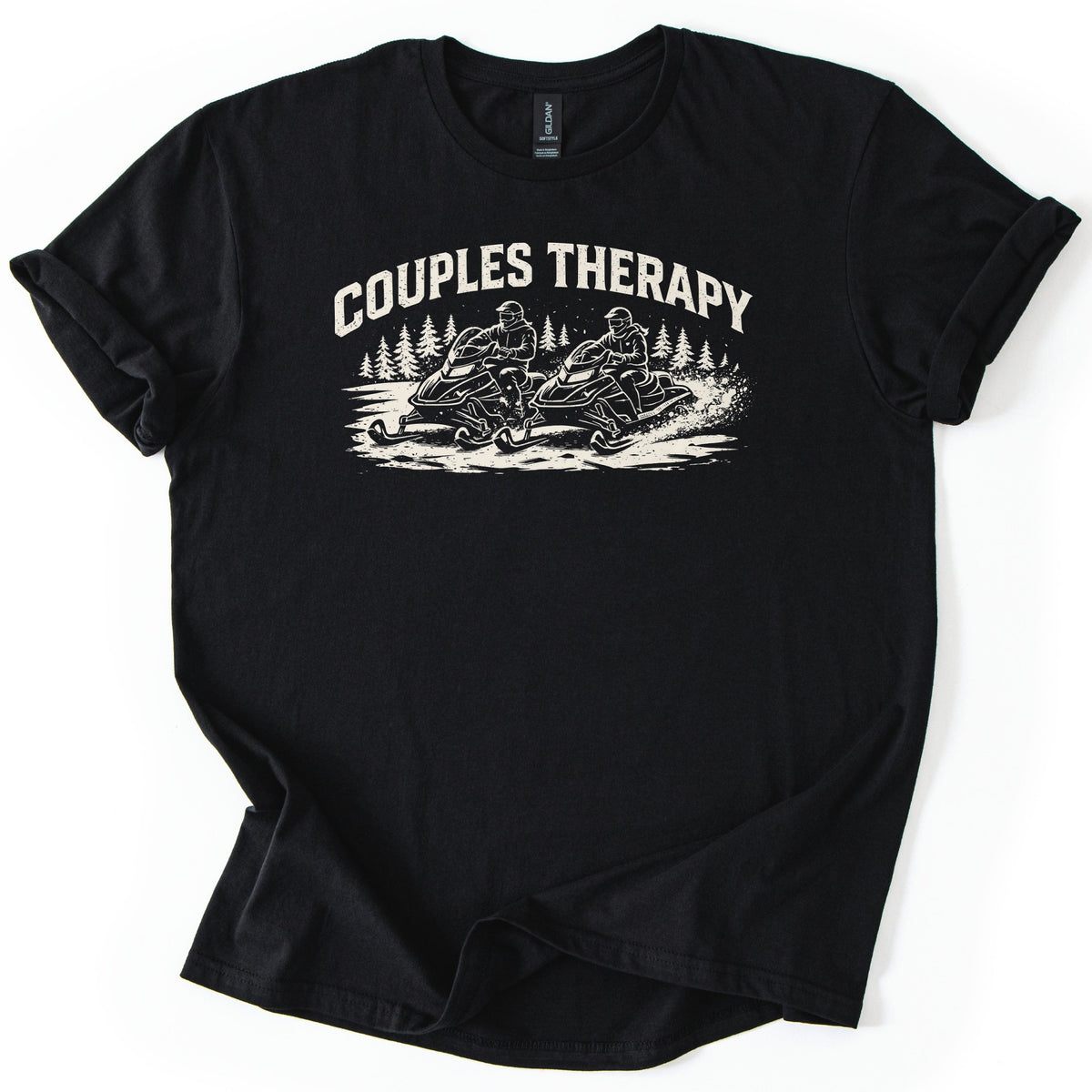 Couples Therapy Snowmobile Comfort Colors T-shirt in Black, laid flat. Cream print of the arched words Couples Therapy above two helmeted riders on snowmobiles racing side by side through snow with pine trees behind. Sleeves are cuffed on a white background.