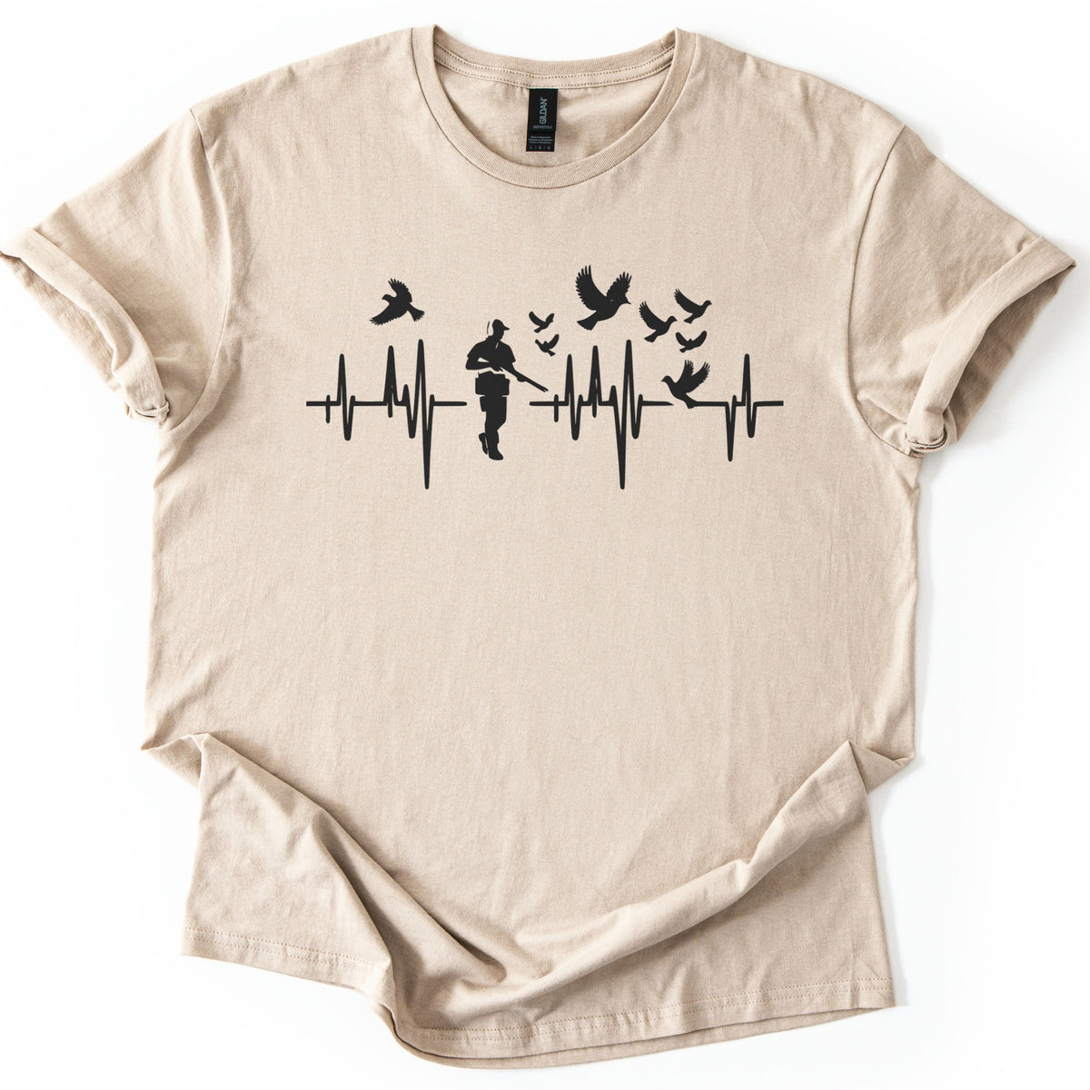 Bird Hunter Comfort Colors T-shirt in Sand, laid flat. A black heartbeat line crosses the chest around a silhouetted hunter in a cap holding a shotgun, with birds flying above and one perched on the line. No words. Cuffed sleeves on a white background.