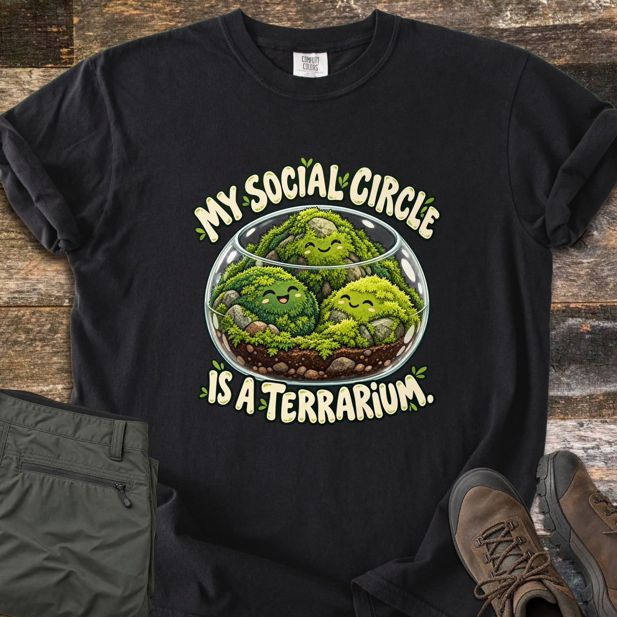 My Social Circle Is a Terrarium Comfort Colors T-shirt in Black, laid flat. A glass bowl terrarium holds three smiling moss balls among rocks and soil, between the words My Social Circle and Is a Terrarium. Folded olive pants and brown hiking boots sit on weathered wood planks.