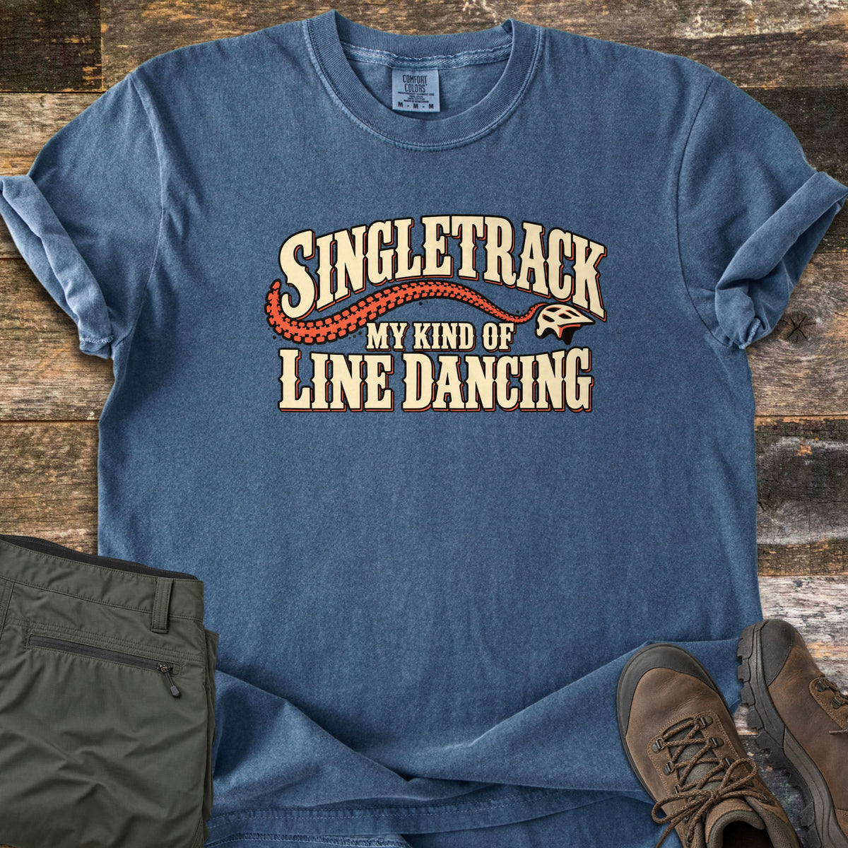 Singletrack My Kind of Line Dancing Comfort Colors T-shirt in Denim, laid flat. Cream western-style lettering reads Singletrack, My Kind of Line Dancing, with an orange tire track curving to a bike helmet. Shown on weathered wood with folded green hiking pants and brown hiking boots.