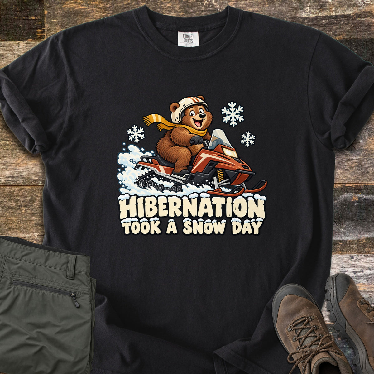 Hibernation Took a Snow Day Comfort Colors T-shirt in Black, laid flat. A cartoon bear in a white helmet and yellow scarf rides a red snowmobile among snowflakes above the words Hibernation Took a Snow Day. Olive hiking pants and brown boots sit on weathered boards.