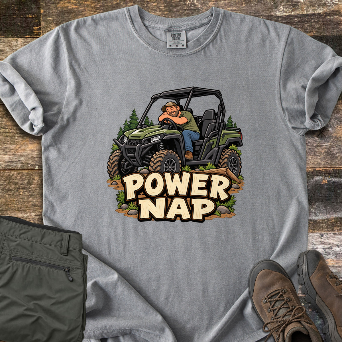 Power Nap Comfort Colors T-shirt in Grey, laid flat. A cartoon man in a cap naps with his head on the steering wheel of a green side-by-side UTV among pine trees, above the words Power Nap. Folded olive pants and brown hiking boots sit on a weathered wood surface.