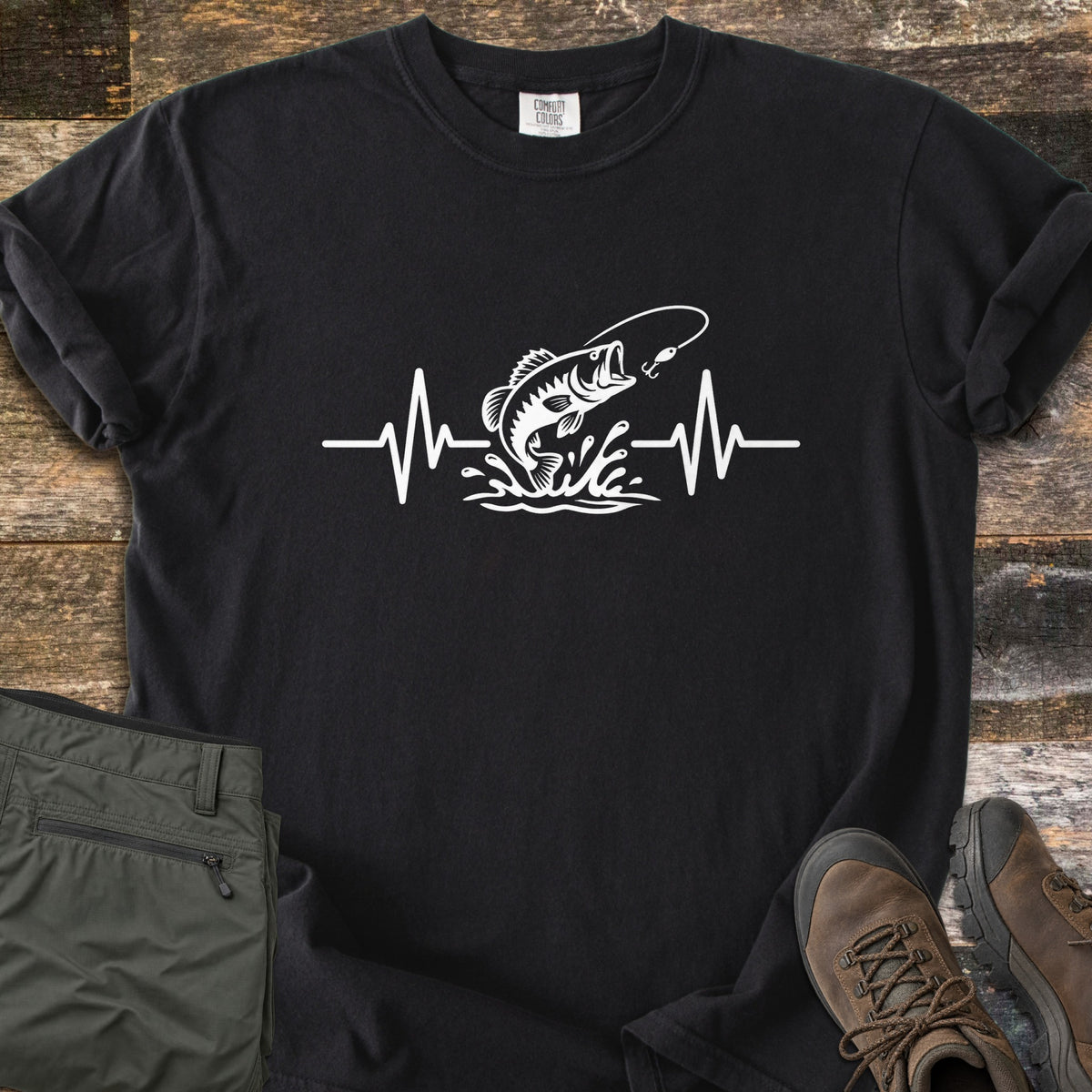 Fishing Heartbeat Comfort Colors T-shirt in Black, laid flat. A white print of a largemouth bass leaping from splashing water toward a fishing lure, set in the middle of a heartbeat line. Sleeves are rolled, on rustic wood with olive cargo shorts and brown hiking boots.