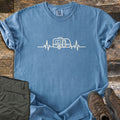 Camper Heartbeat Comfort Colors T-shirt in Blue Jean, laid flat. A cream line drawing of a teardrop camper trailer sits in the middle of a heartbeat line across the chest. Folded olive pants and brown hiking boots rest on a rustic wood background.