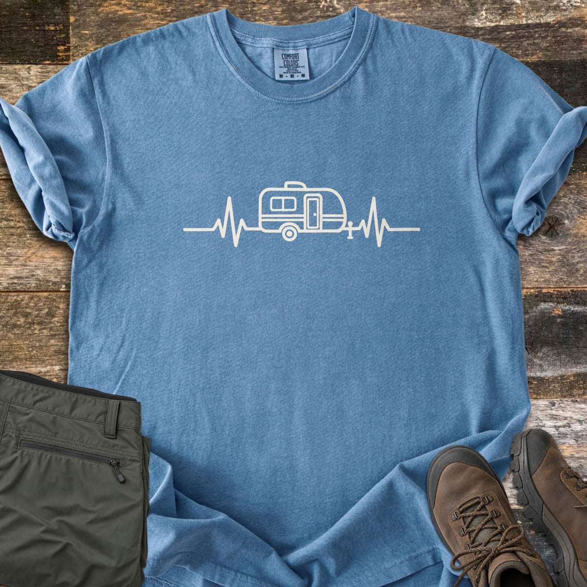 Camper Heartbeat Comfort Colors T-shirt in Blue Jean, laid flat. A cream line drawing of a teardrop camper trailer sits in the middle of a heartbeat line across the chest. Folded olive pants and brown hiking boots rest on a rustic wood background.