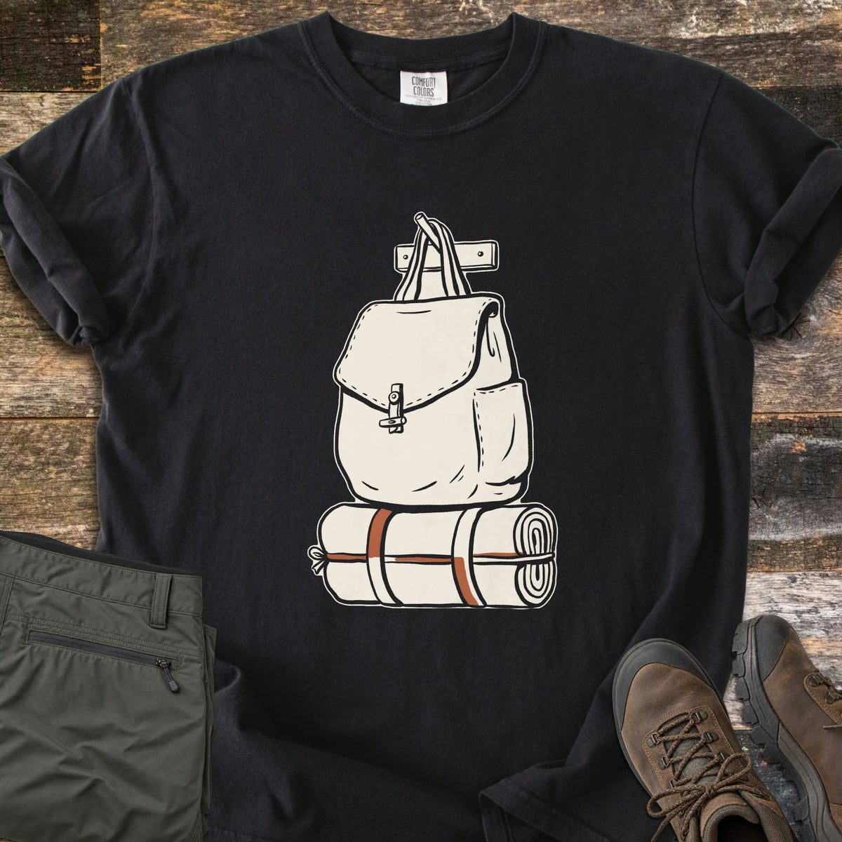 Backpack And Bedroll Comfort Colors T-shirt in Black, laid flat. A cream canvas backpack hanging from a wall peg above a bedroll tied with rust-orange straps. Olive hiking pants and brown hiking boots sit beside it on weathered wood planks.