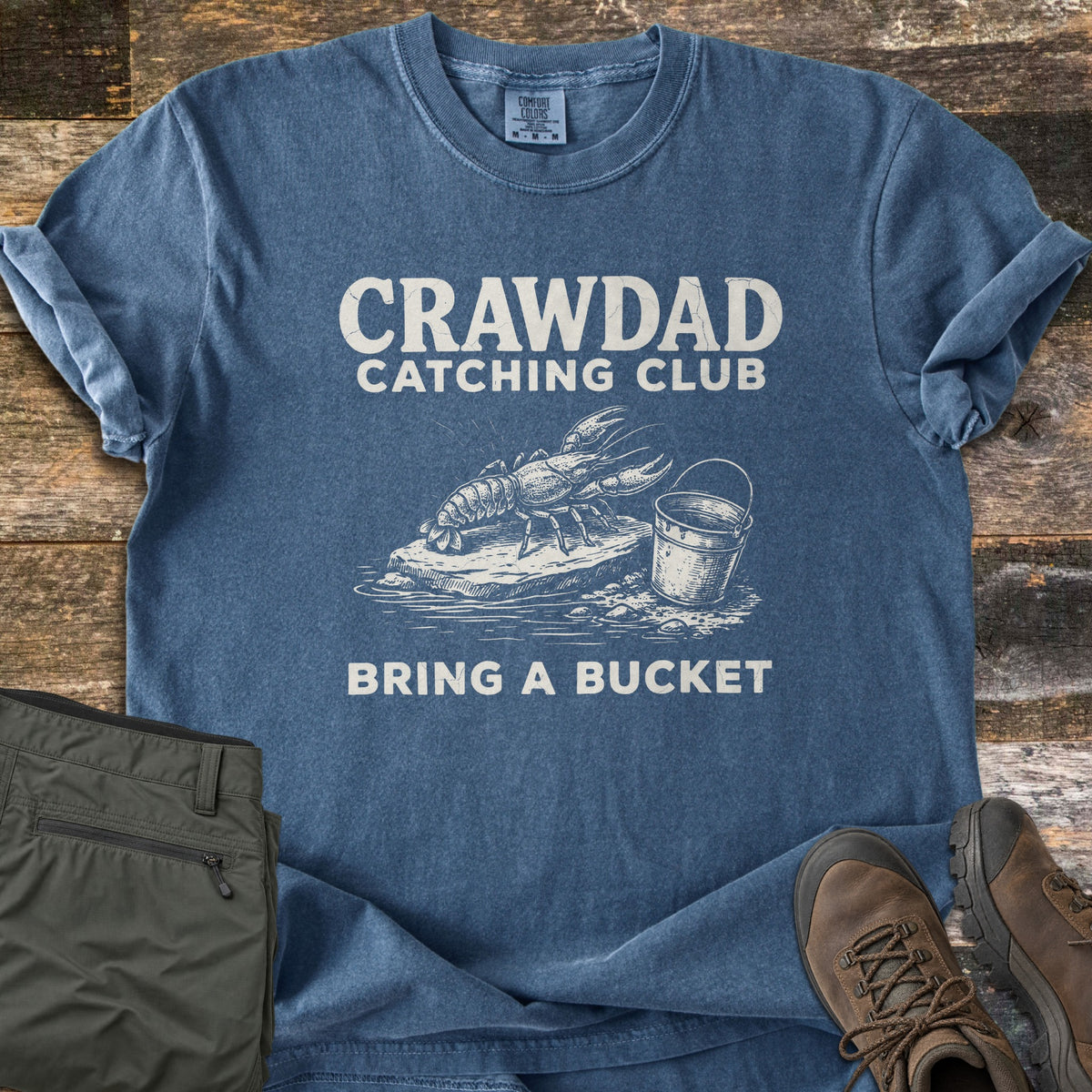 Crawdad Catching Club Comfort Colors T-shirt in Denim, laid flat. An etched-style crawdad on a flat rock by a metal bucket at the water's edge, between the words Crawdad Catching Club and Bring a Bucket. Shown on weathered wood with olive pants and brown hiking boots.