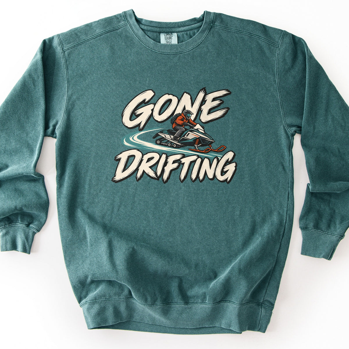 Gone Drifting Comfort Colors crewneck sweatshirt in Blue Spruce, laid flat. The words Gone Drifting in cream brush lettering surround a rider in an orange jacket and helmet on a snowmobile kicking up a teal snow trail. It lies on a white background.