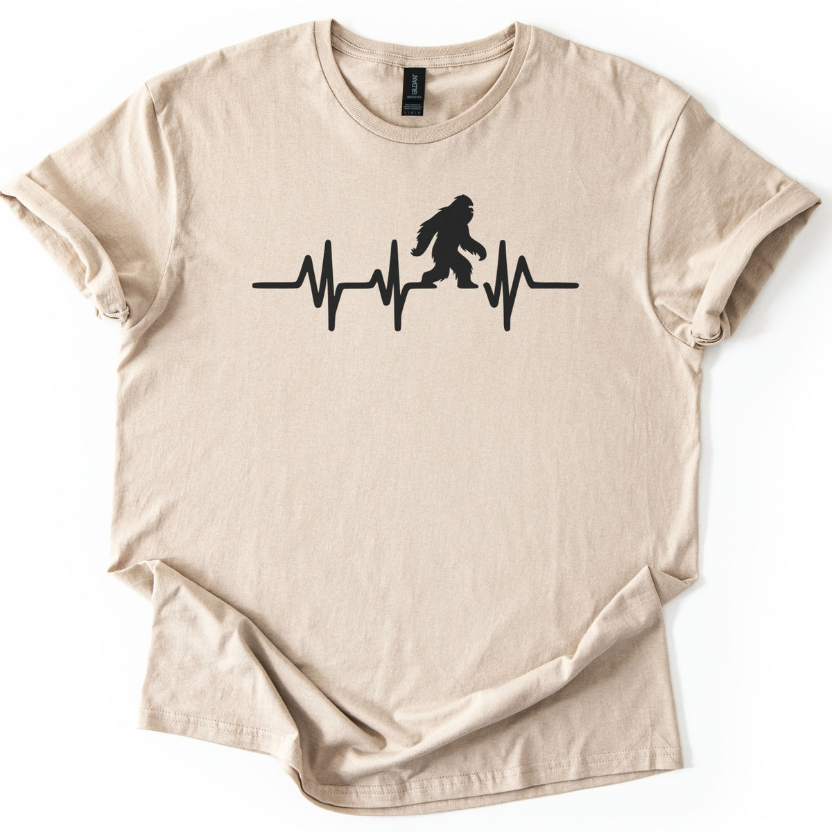 Yeti Heartbeat Comfort Colors T-shirt in Sand, laid flat. A black heartbeat line runs across the chest, with a walking Bigfoot silhouette standing within it; there are no words. The sleeves are cuffed and the shirt lies on a white background.