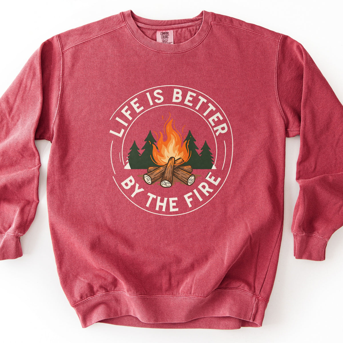 Life Is Better By The Fire Ring Comfort Colors crewneck sweatshirt in Crimson, laid flat. A circular badge shows a campfire of crossed logs with orange flames before dark green pine trees, ringed by the words Life Is Better By The Fire in cream lettering. It lies on a white surface.