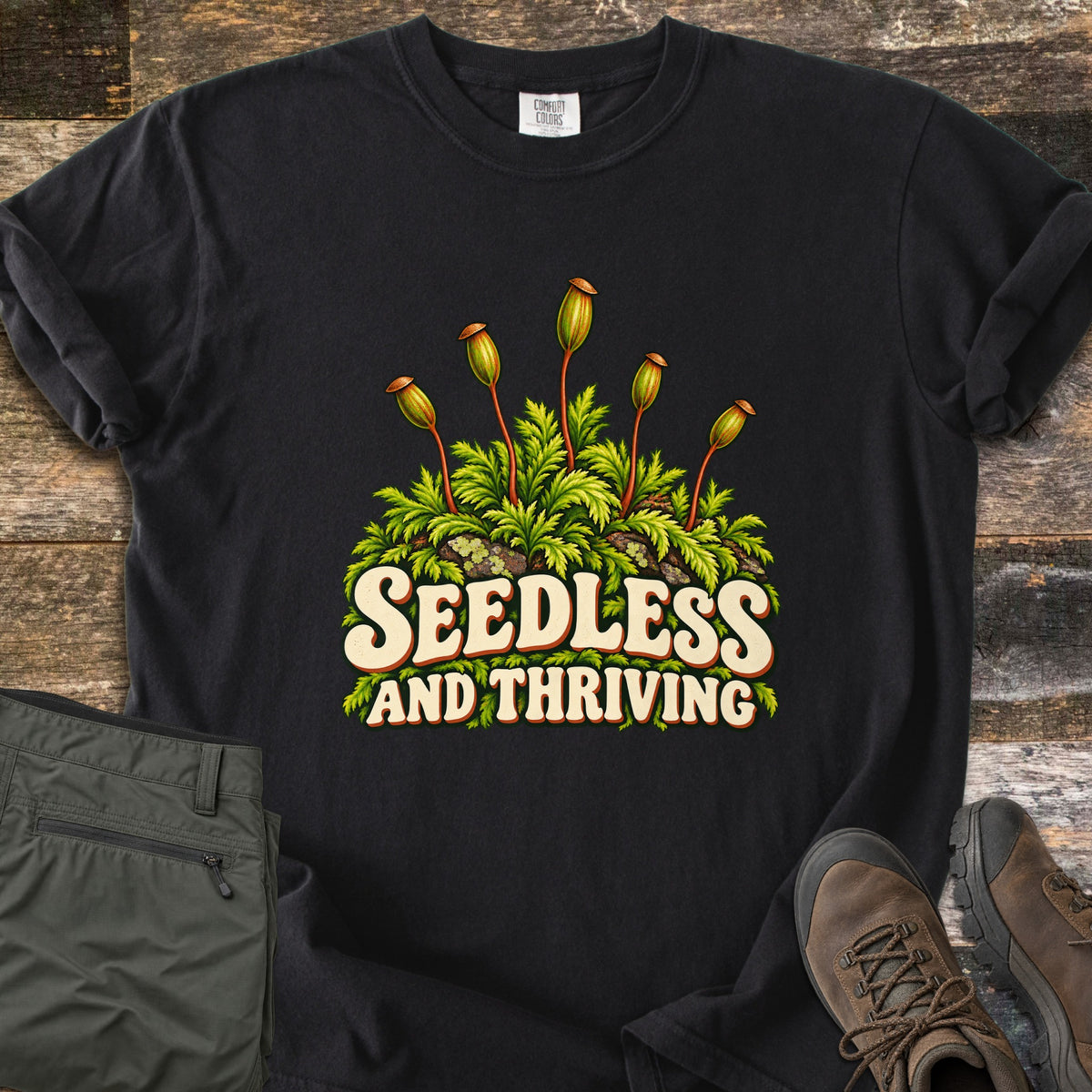 Seedless and Thriving Comfort Colors T-shirt in Black, laid flat. Green moss over mossy rocks with tall stalks topped by spore capsules sits above the words Seedless and Thriving in cream retro letters. It lies on weathered wood planks next to olive hiking pants and brown boots.