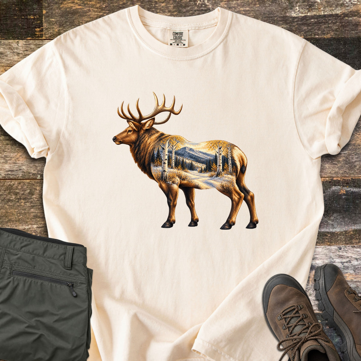 Aspen Pass Elk Comfort Colors T-shirt in Ivory, laid flat. A bull elk in profile whose body holds a snowy mountain landscape with golden aspen trees and pines. Laid on dark weathered wood next to folded olive hiking pants and brown leather hiking boots.