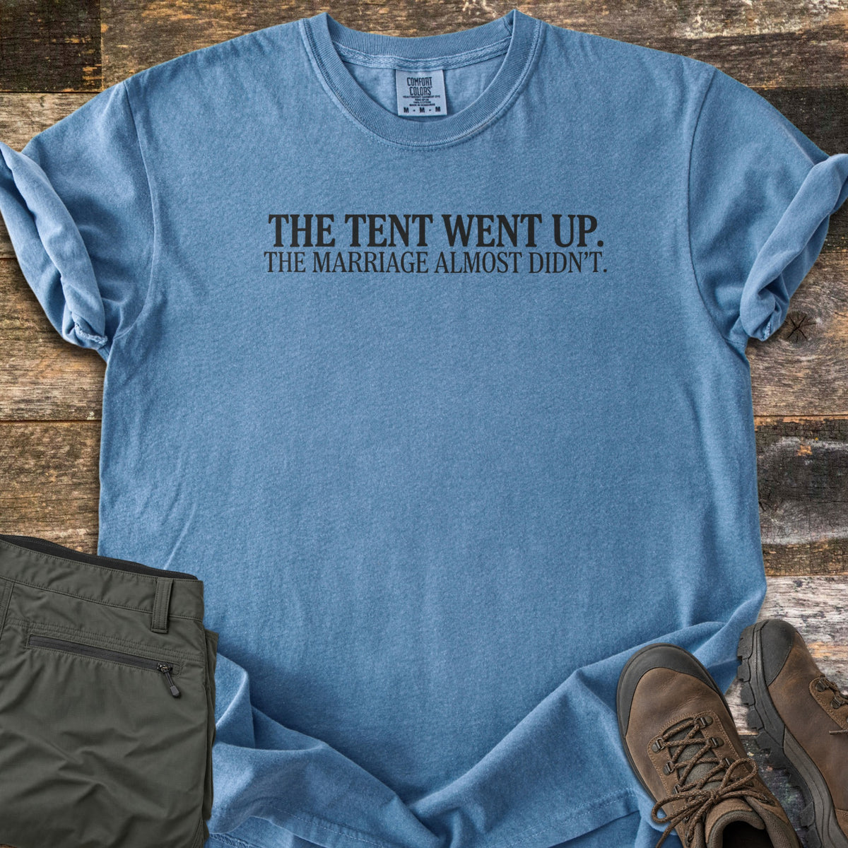 The Tent Went Up Comfort Colors T-shirt in Blue Jean, laid flat. Black serif text across the chest reads The Tent Went Up. The Marriage Almost Didn't. Sleeves are rolled, with folded olive hiking pants and brown hiking boots beside it on weathered wood planks.