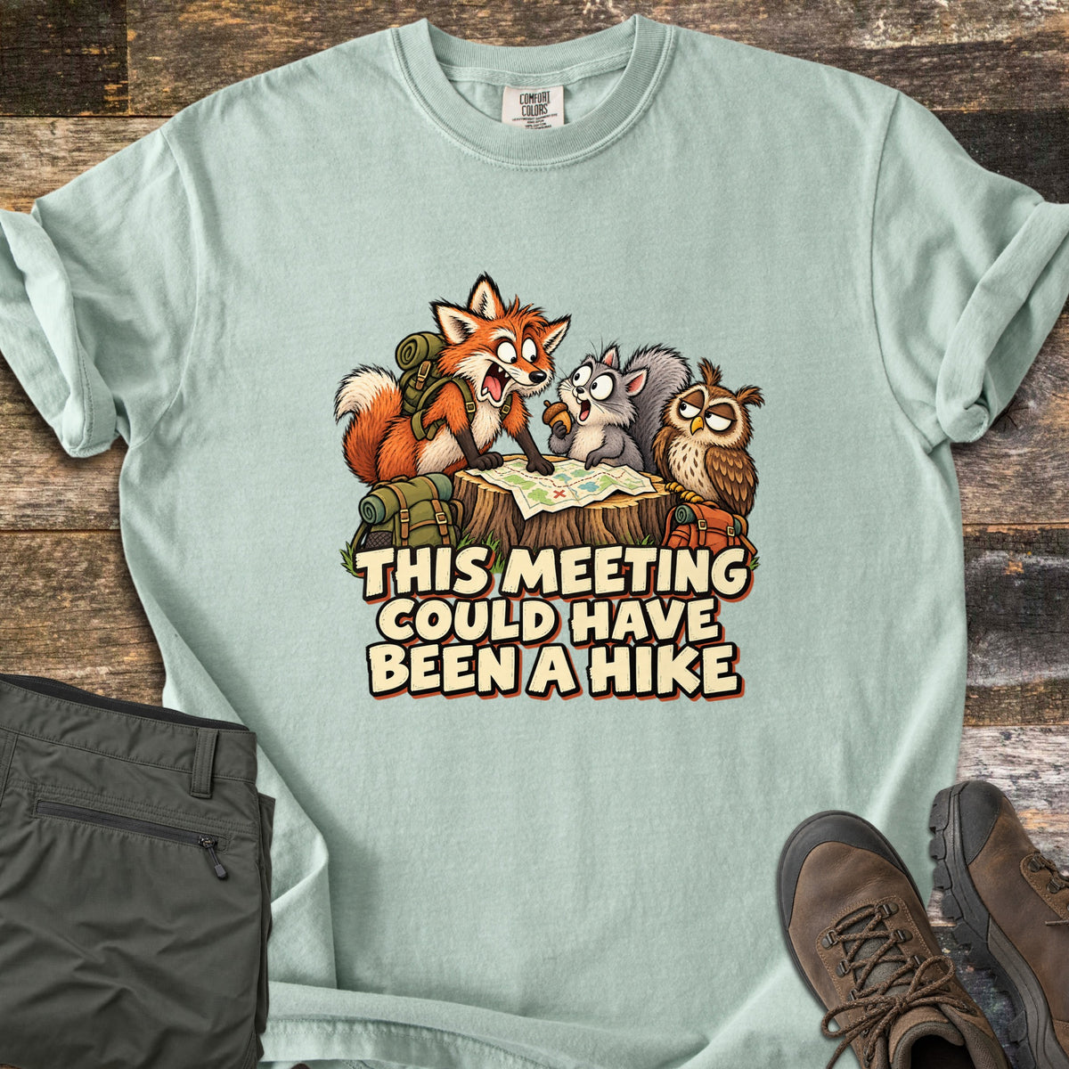 This Meeting Could Have Been a Hike Comfort Colors T-shirt in Bay, laid flat. A cartoon fox, squirrel and owl with hiking packs gather at a trail map spread on a stump above the words This Meeting Could Have Been a Hike. Folded olive hiking pants and brown boots rest on rustic wood.
