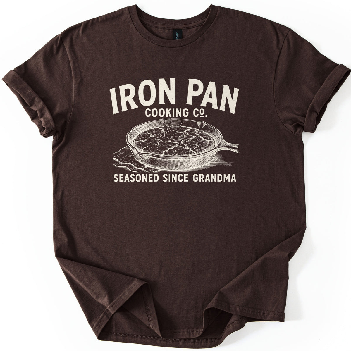 Iron Pan Cooking Co Comfort Colors T-shirt in Dark Chocolate, laid flat. Cream print of the words Iron Pan Cooking Co. above an engraved-style cast iron skillet of cornbread topped with melting butter on a cloth, with Seasoned Since Grandma below. Cuffed sleeves on a white background.