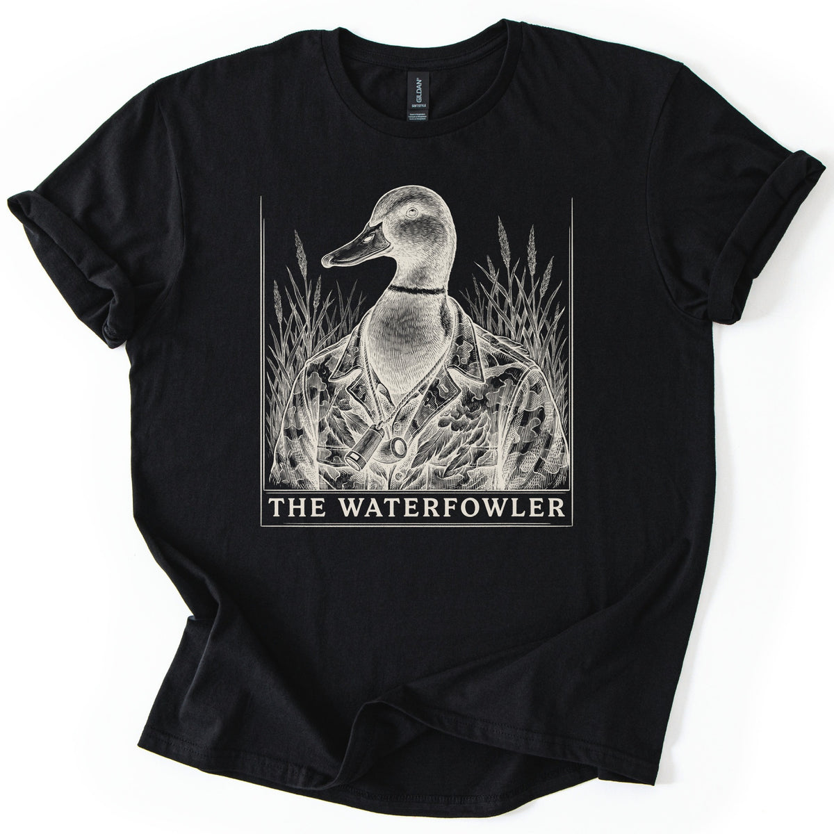 The Waterfowler Comfort Colors T-shirt in Black, laid flat. A cream engraving-style drawing of a mallard duck in a camo shirt with a duck call on a lanyard, set in tall marsh grass inside a thin frame, above the words THE WATERFOWLER. Sleeves cuffed, on white.