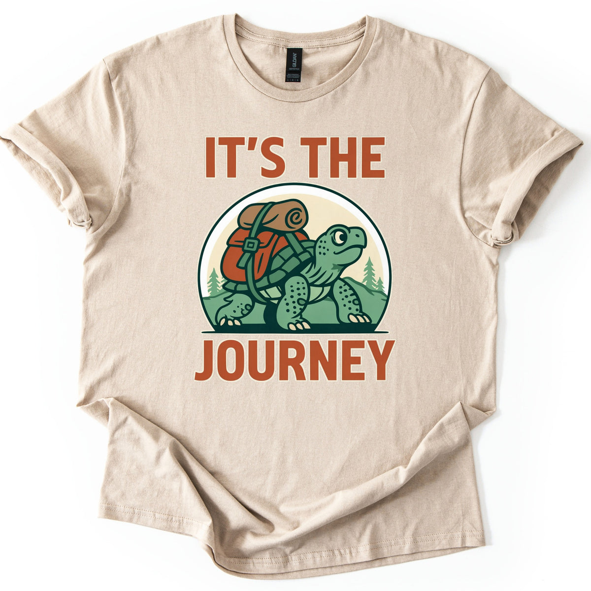 Slow Turtle Hiker Journey Comfort Colors T-shirt in Sand, laid flat. A green cartoon turtle carrying a red backpack with a rolled sleeping bag walks past pine trees, framed by the words It's the Journey in rust-orange letters. Sleeves are cuffed on a white background.