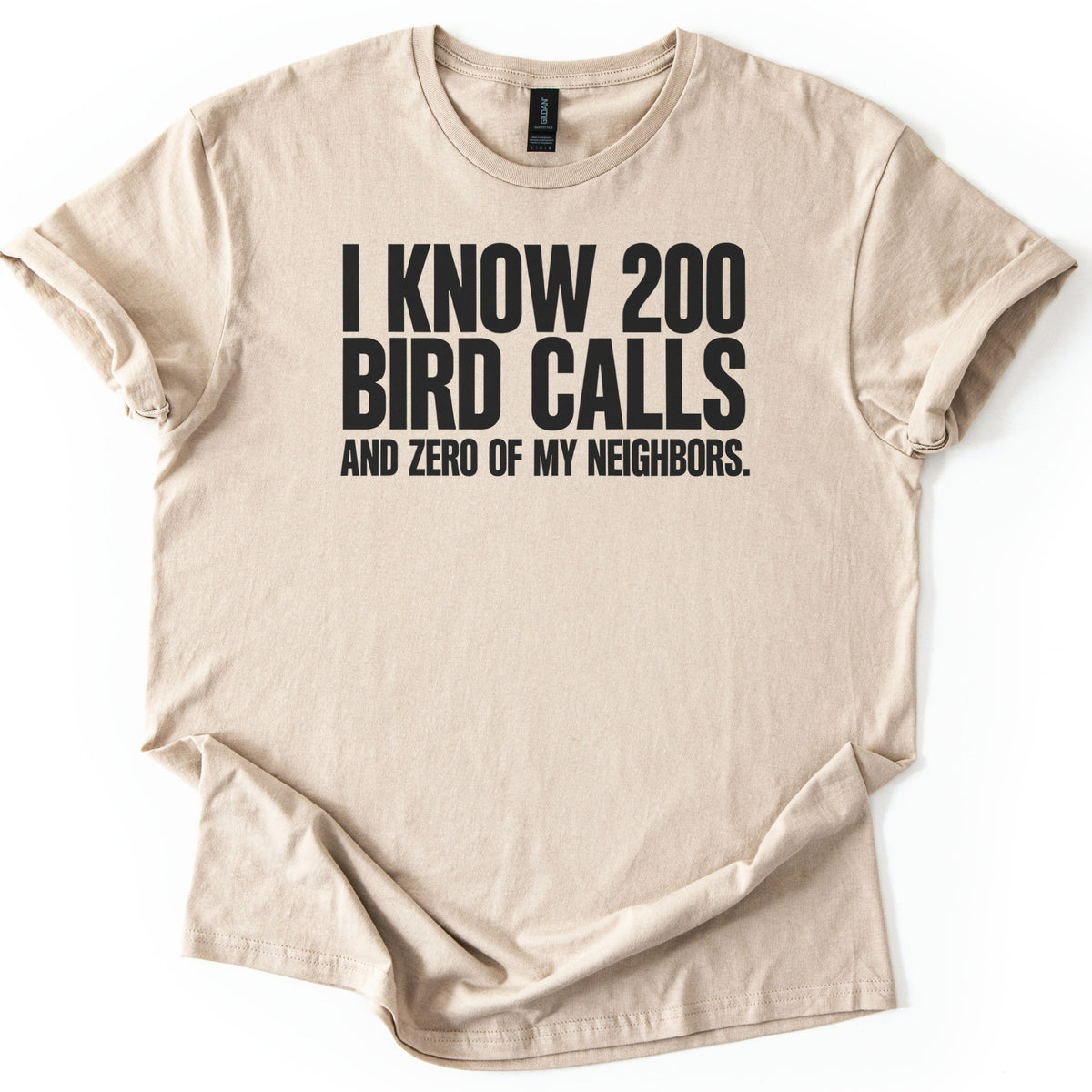 I Know 200 Bird Calls and Zero of My Neighbors Comfort Colors T-shirt in Sand, laid flat. Bold black capital letters across the chest read I Know 200 Bird Calls and Zero of My Neighbors. The shirt lies on a white background with rolled sleeves and a loosely rumpled hem.