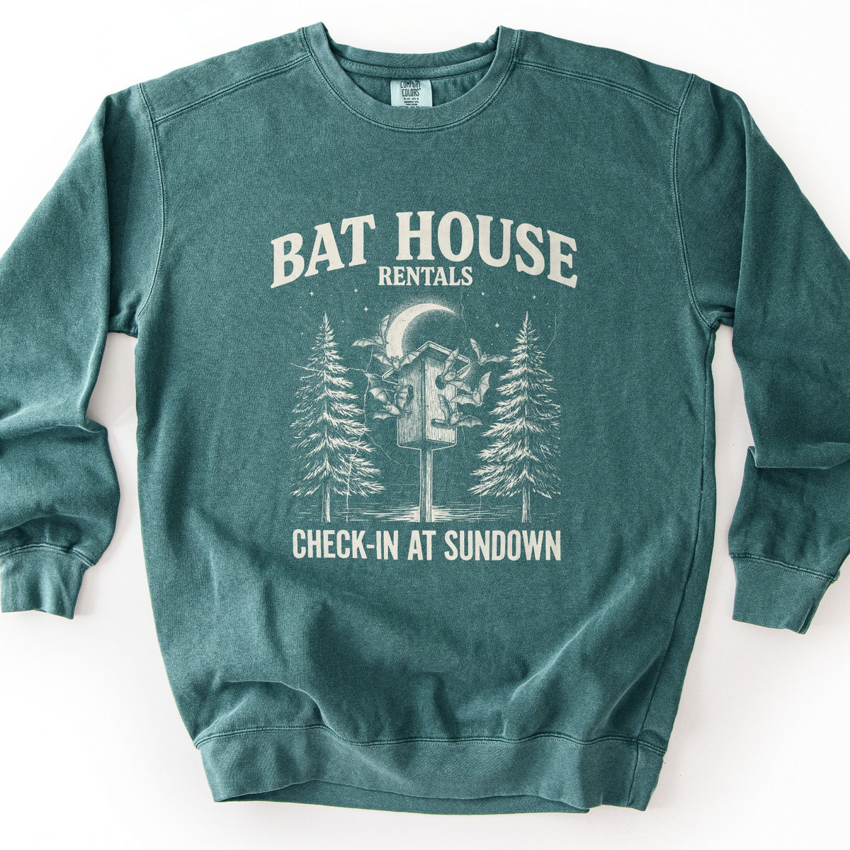 Bat House Rentals Comfort Colors crewneck sweatshirt in Blue Spruce, laid flat. Cream print of a wooden bat house on a post with bats flying around it, a crescent moon, stars and two pine trees, under the words Bat House Rentals and above Check-In at Sundown. Shown on a white background.