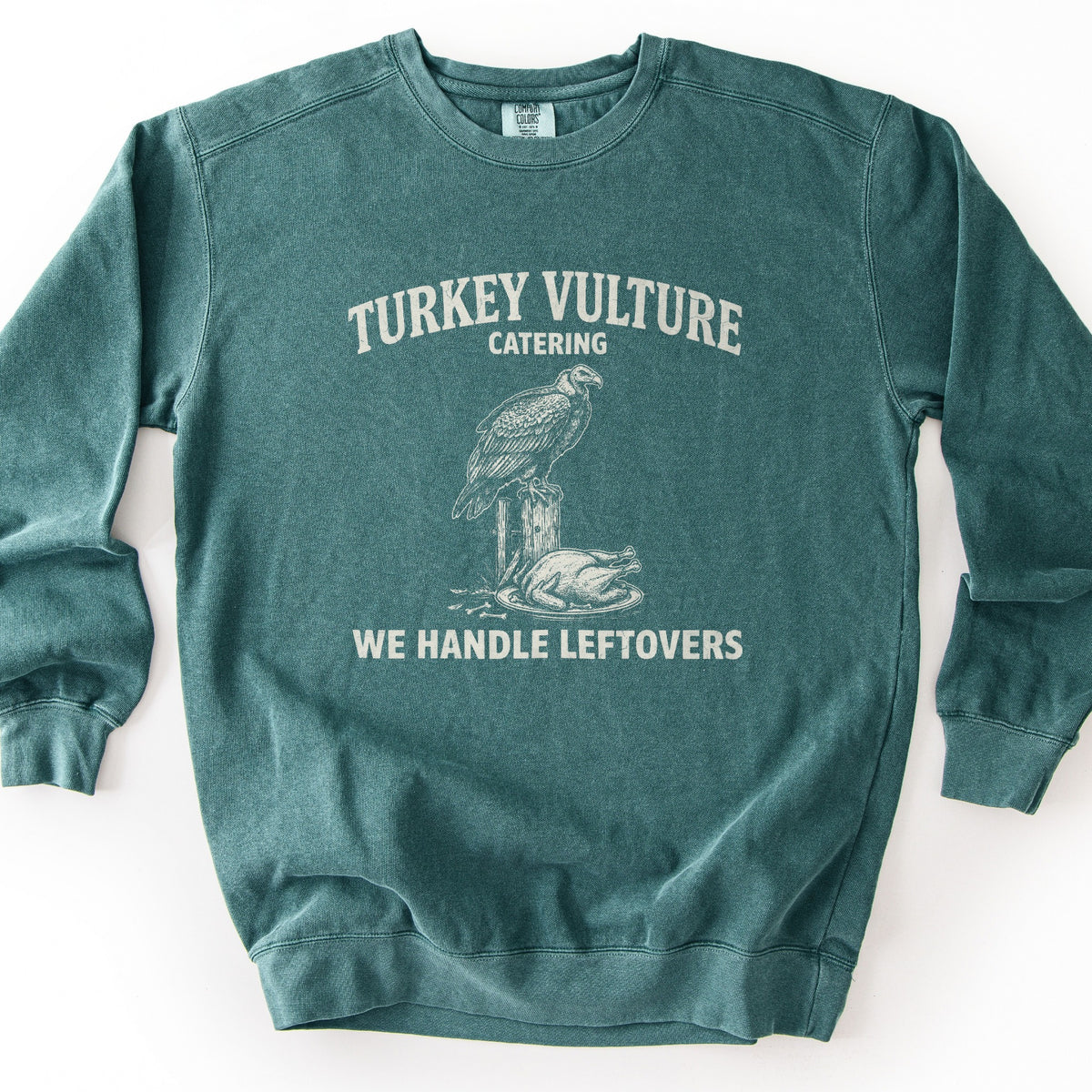 Turkey Vulture Catering Comfort Colors crewneck sweatshirt in Blue Spruce, laid flat. Cream print of a vulture perched on a wooden post beside a roasted turkey on a platter, with the words Turkey Vulture Catering above and We Handle Leftovers below. Shown on a plain white background.