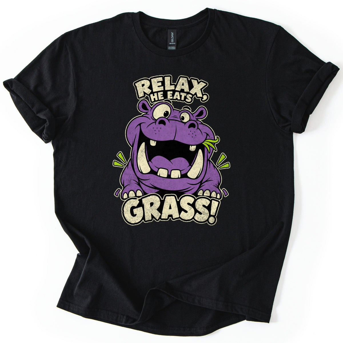 Relax He Eats Grass Comfort Colors T-shirt in Black, laid flat. A grinning purple cartoon hippo with big tusks and a blade of grass in its mouth sits between the words RELAX, HE EATS and GRASS! in cream letters. The shirt lies on white with rolled sleeves.