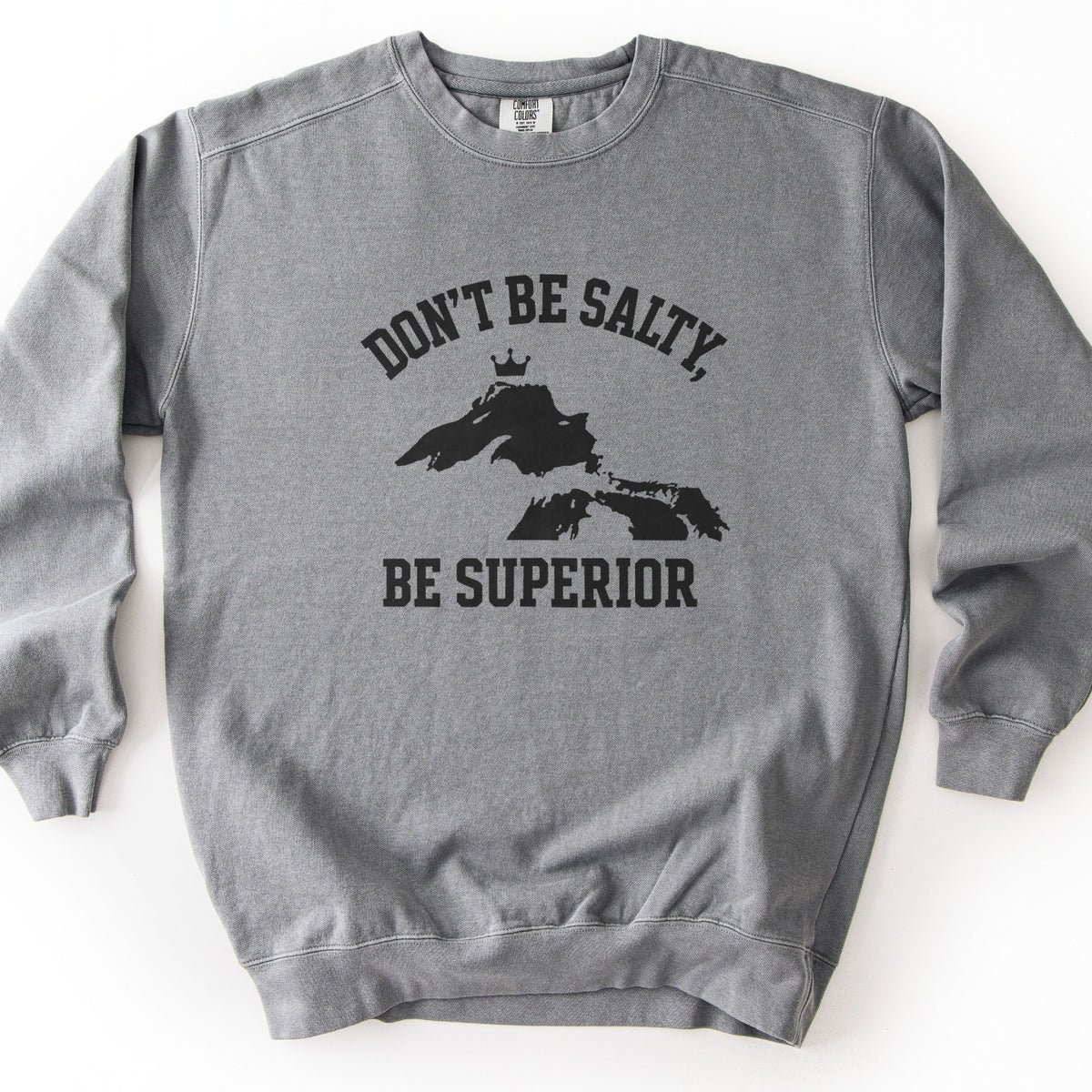 Be Superior Comfort Colors crewneck sweatshirt in Grey, laid flat. Black print of the words Don't Be Salty, arched above a silhouette of Lake Superior topped with a small crown, with Be Superior below. Laid on a white background.
