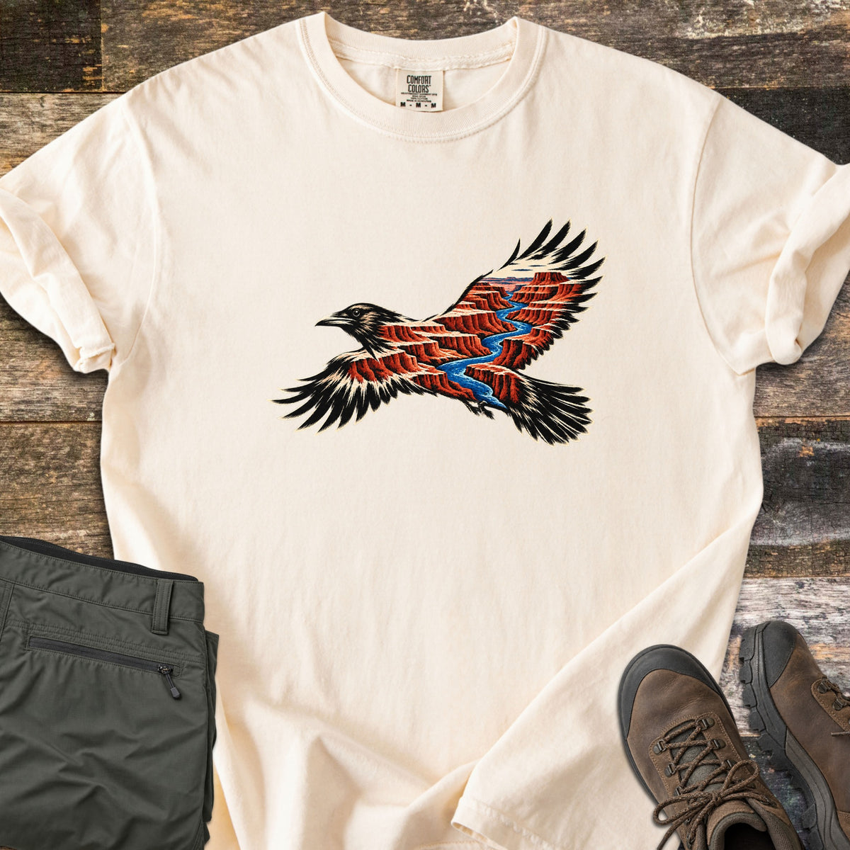 Red Canyon Raven Comfort Colors T-shirt in Ivory, laid flat. A soaring black raven, its body showing red canyon cliffs and a winding blue river. The cream shirt has rolled sleeves and lies on weathered wood with folded olive hiking pants and brown boots.