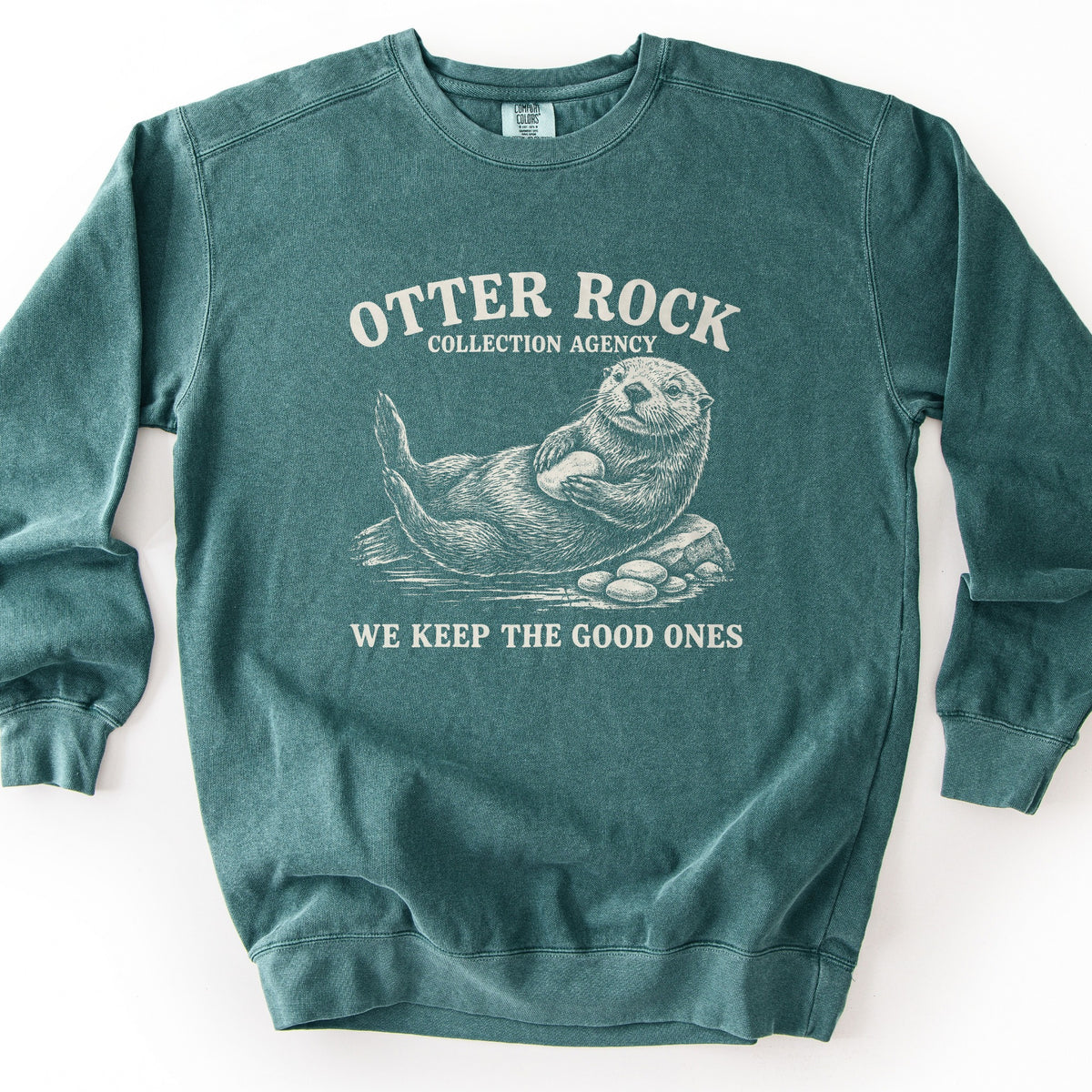 Otter Rock Collection Agency Comfort Colors crewneck sweatshirt in Blue Spruce, laid flat. A cream sketch-style sea otter floats on its back hugging a smooth stone beside a rock and pebbles, under the words Otter Rock Collection Agency and above We Keep The Good Ones. Shown on a white background.