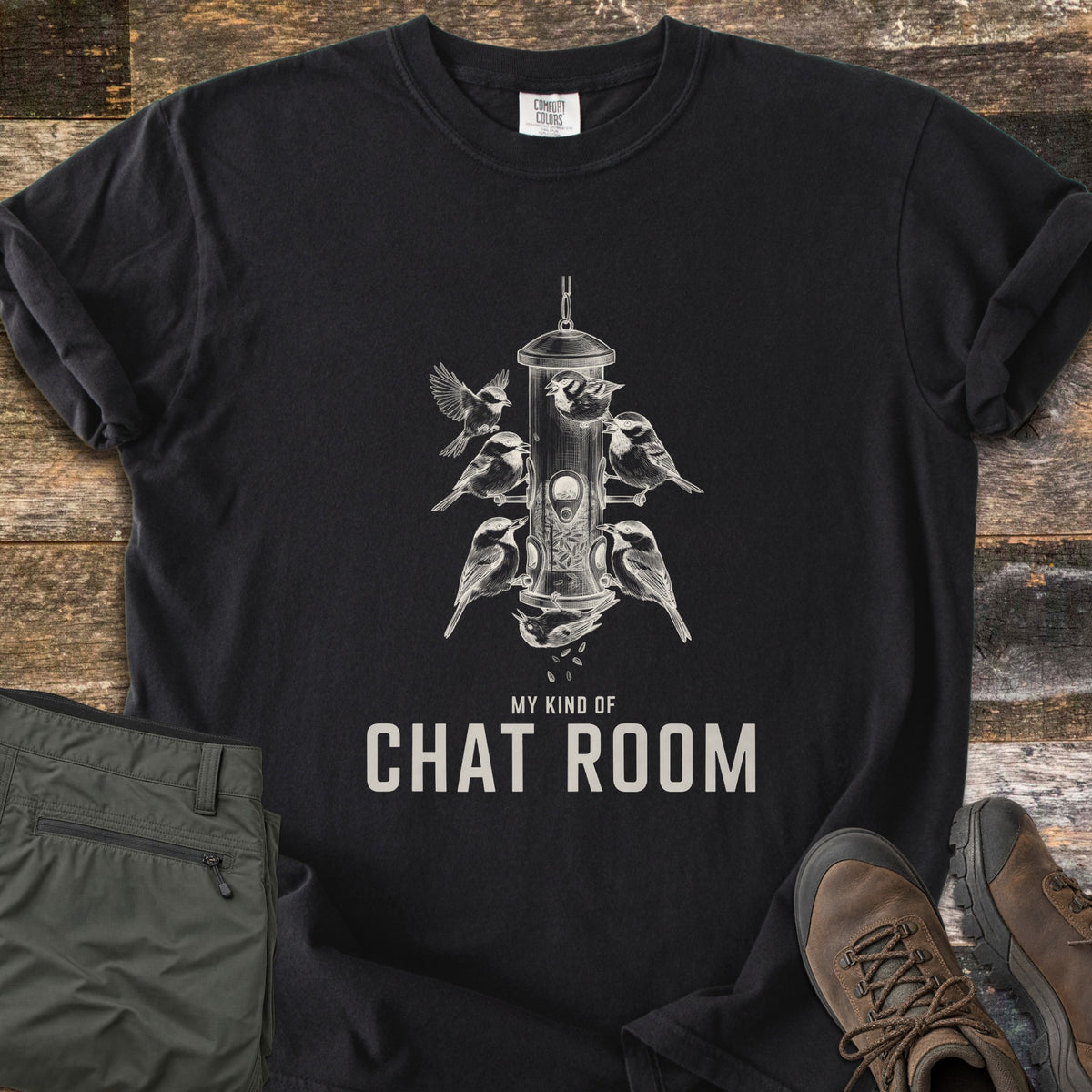 My Kind Of Chat Room Comfort Colors T-shirt in Black, laid flat. A cream sketch of chickadees perched around a hanging tube bird feeder, one upside down below it, above the words My Kind Of Chat Room. Olive hiking pants and brown boots lie beside it on weathered wood.