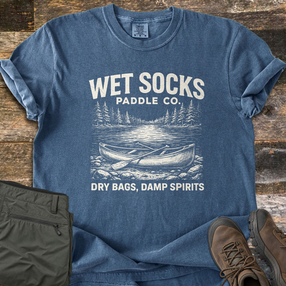 Wet Socks Paddle Co Comfort Colors T-shirt in Denim, laid flat. An etched-style canoe with a paddle rests on a rocky shore by a pine-lined lake, under the words Wet Socks Paddle Co. and above Dry Bags, Damp Spirits. Olive pants and brown hiking boots sit on weathered wood.