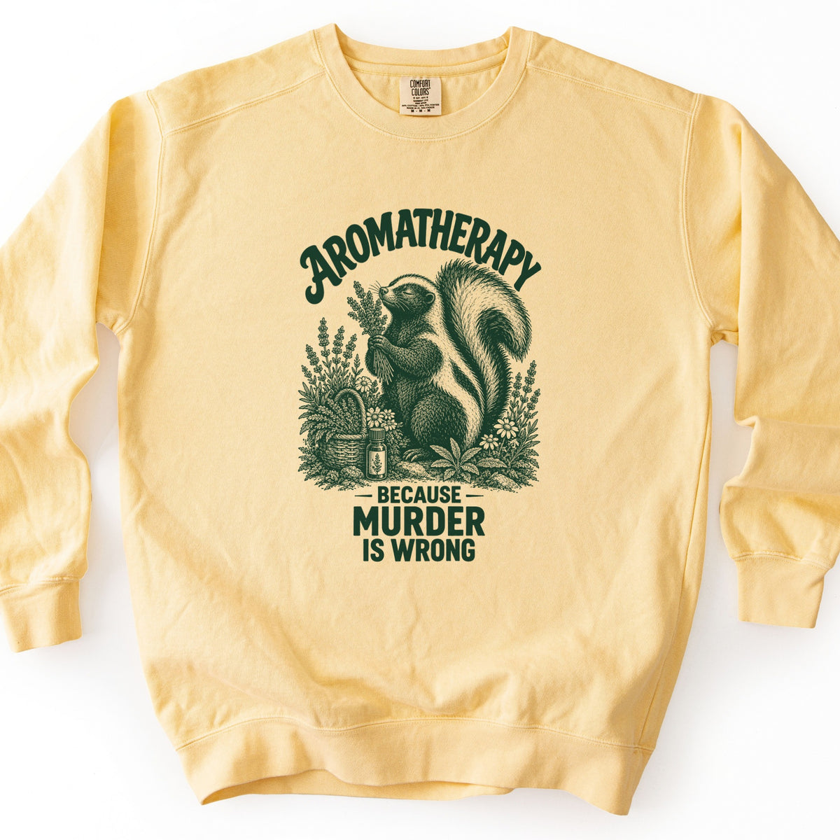 Aromatherapy Because Murder Is Wrong Comfort Colors crewneck sweatshirt in Butter, laid flat. Dark green line art of a skunk sniffing a bundle of lavender among wildflowers, a basket and an essential oil bottle, with Aromatherapy above and Because Murder Is Wrong below. Shown on a white surface.