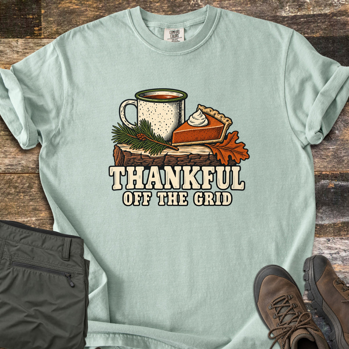 Thankful Off the Grid Comfort Colors T-shirt in Bay, laid flat. A speckled enamel camp mug and a slice of pumpkin pie with whipped cream sit on a log with a pine sprig and an oak leaf above the words Thankful Off the Grid. Olive hiking pants and brown boots lie on weathered wood.