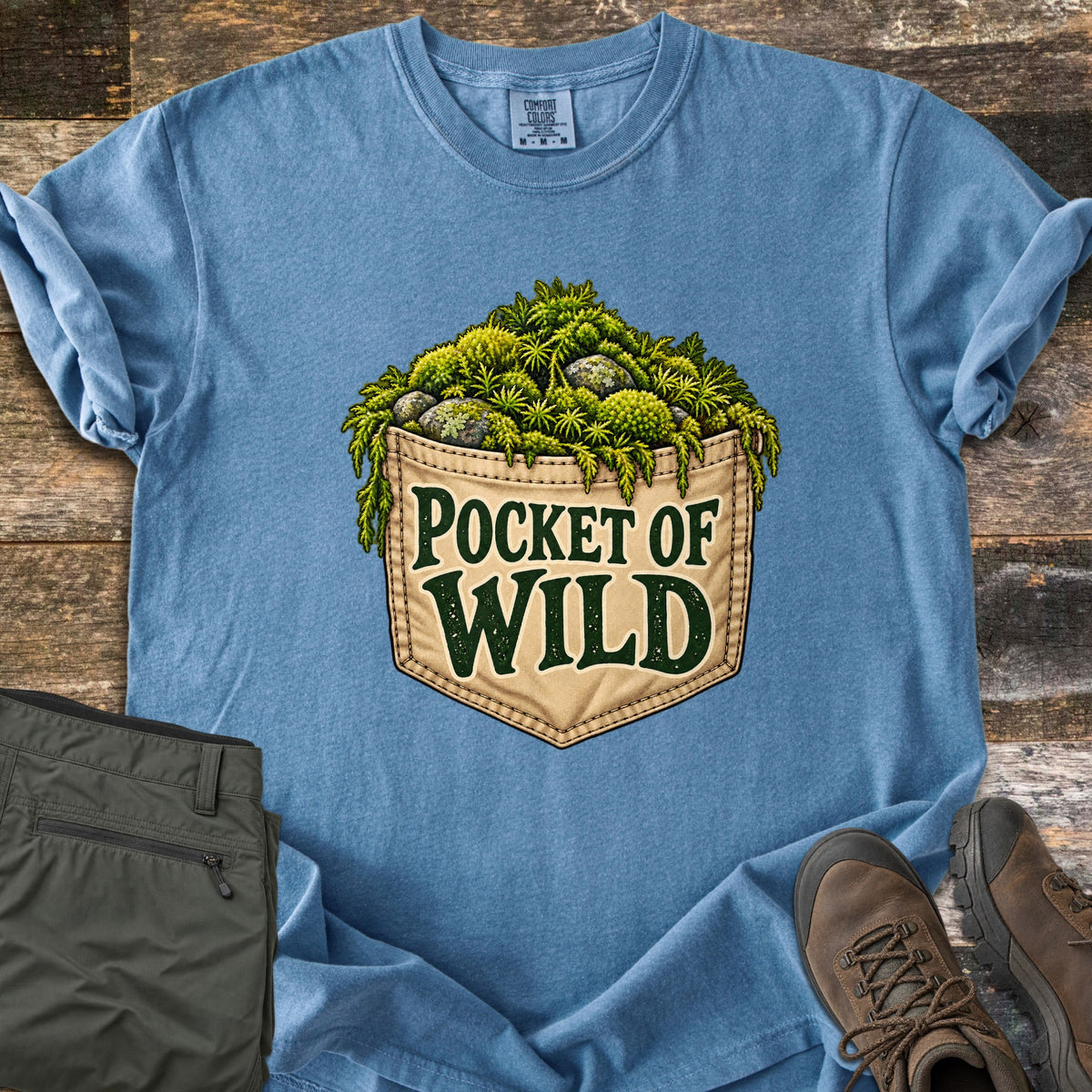 Pocket of Wild Comfort Colors T-shirt in Blue Jean, laid flat. A tan stitched jeans pocket overflowing with green moss and lichen-covered stones, printed with the words Pocket of Wild. Shown on weathered wood with folded olive hiking pants and brown hiking boots.