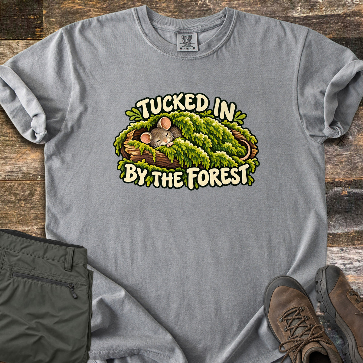 Tucked in by the Forest Comfort Colors T-shirt in Grey, laid flat. A sleeping cartoon mouse lies under a blanket of green moss in a hollow log between the words Tucked In By The Forest. Folded olive hiking pants and brown hiking boots sit on weathered wood planks.