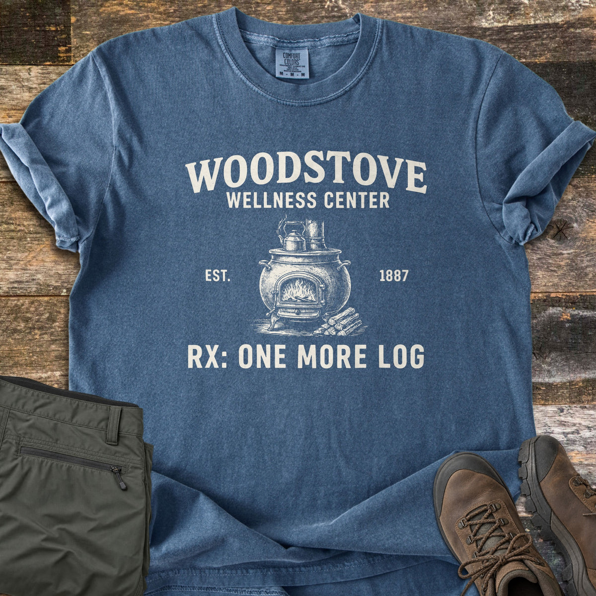 Woodstove Wellness Center Comfort Colors T-shirt in Denim, laid flat. A sketched wood stove with a kettle, fire and logs sits between EST. and 1887 under the words Woodstove Wellness Center, with Rx: One More Log below. Olive pants and brown hiking boots rest on weathered wood planks.