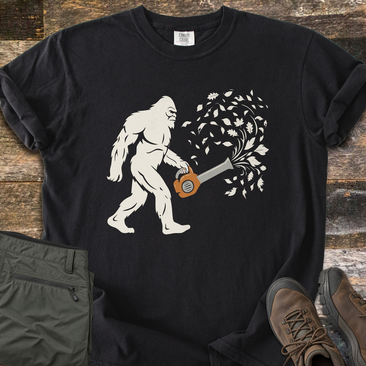 Bigfoot Leaf Blower Comfort Colors T-shirt in Black, laid flat. A white Bigfoot silhouette walks while holding an orange leaf blower that sends a swirl of white leaves into the air. Folded olive pants and brown hiking boots sit beside it on weathered wood boards.